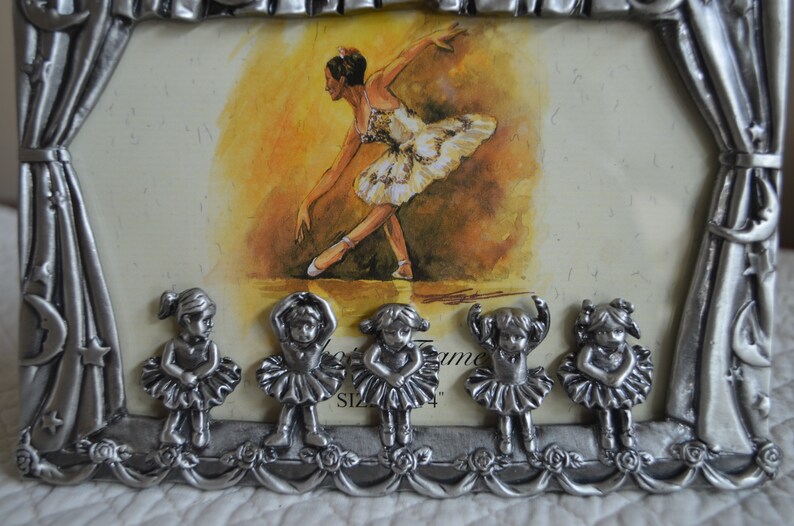 May include: A silver-tone picture frame with a ballerina image inside. The frame has a decorative design with a curtain and stars. Five small ballerina figurines are attached to the bottom of the frame.