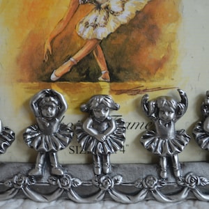 May include: Five silver ballerina figurines with white tutus and black hair. The figurines are arranged in a row on a silver frame with floral accents. The frame has the text "Picture Frame Size 4" printed on it.