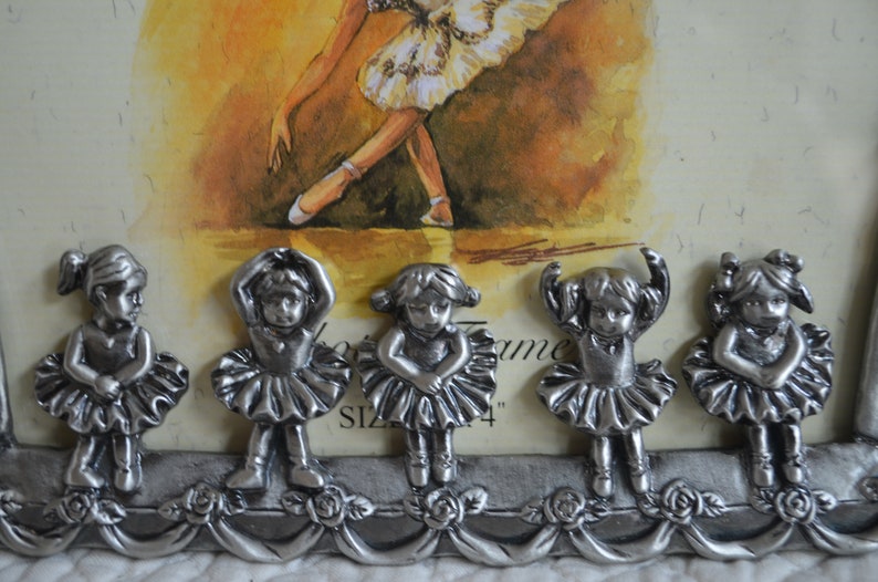 May include: Five silver ballerina figurines with white tutus and black hair. The figurines are arranged in a row on a silver frame with floral accents. The frame has the text "Picture Frame Size 4" printed on it.