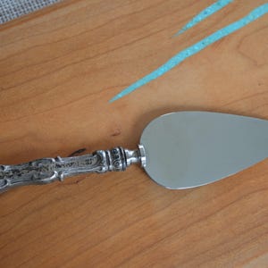 May include: A silver cake server with an ornate handle rests on a wooden cutting board. The server's blade is a smooth, polished silver. The wooden board has a light brown color with streaks of blue paint.