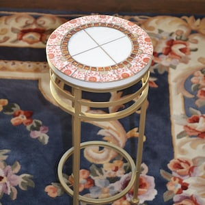 May include: A decorative side table with a round, tiled top featuring a floral and geometric pattern in shades of red, orange, and brown. The table has a gold-colored metal frame and measures approximately 20 inches in height.