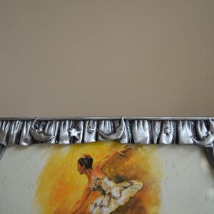 May include: A silver picture frame with a ballerina in a white tutu dancing on a yellow stage. The frame has a decorative border with stars and crescent moons.