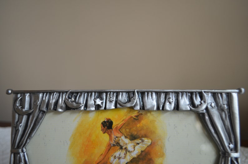 May include: A silver picture frame with a ballerina in a white tutu dancing on a yellow stage. The frame has a decorative border with stars and crescent moons.
