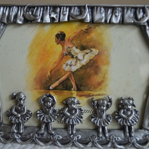May include: A silver-tone picture frame with a ballerina image and five smaller ballerina figurines at the bottom. The frame is decorated with a curtain design and stars.