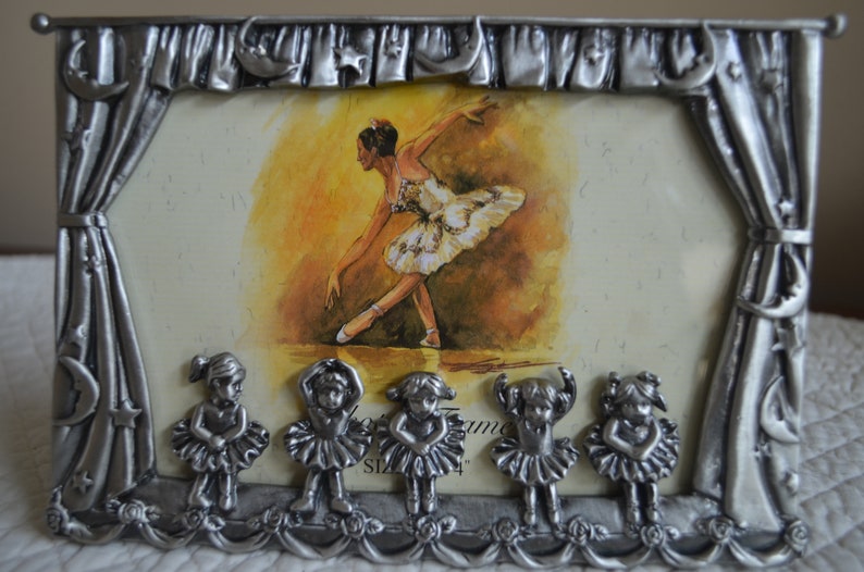 May include: A silver-tone picture frame with a ballerina image and five smaller ballerina figurines at the bottom. The frame is decorated with a curtain design and stars.