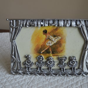 May include: Silver-toned picture frame with a ballerina image and a stage curtain design. The frame features six small ballerina figurines at the bottom.