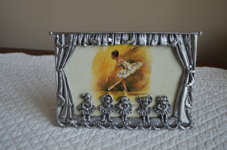 May include: Silver-toned picture frame with a ballerina image and a stage curtain design. The frame features six small ballerina figurines at the bottom.