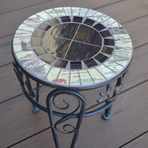 May include: A small round table with a mosaic top made from wine bottle labels. The table has a black metal frame with a decorative scroll design.