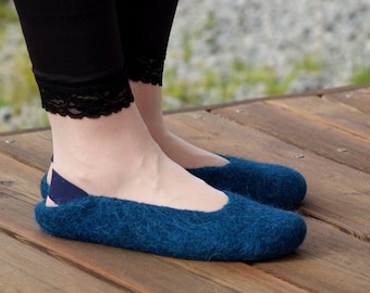 Puede incluir: Un par de zapatillas de lana afieltrada azules. Las zapatillas tienen una puntera redondeada y un pequeño detalle de correa en el talón. Las zapatillas están sobre una superficie de madera.