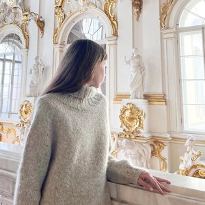 Puede incluir: Un jersey de cuello alto de color gris claro y de gran tama&ntilde;o. El jersey tiene cuello y pu&ntilde;os acanalados. La persona est&aacute; de pie en una habitaci&oacute;n con paredes blancas, detalles dorados y estatuas. La persona est&aacute; mirando por una ventana.
