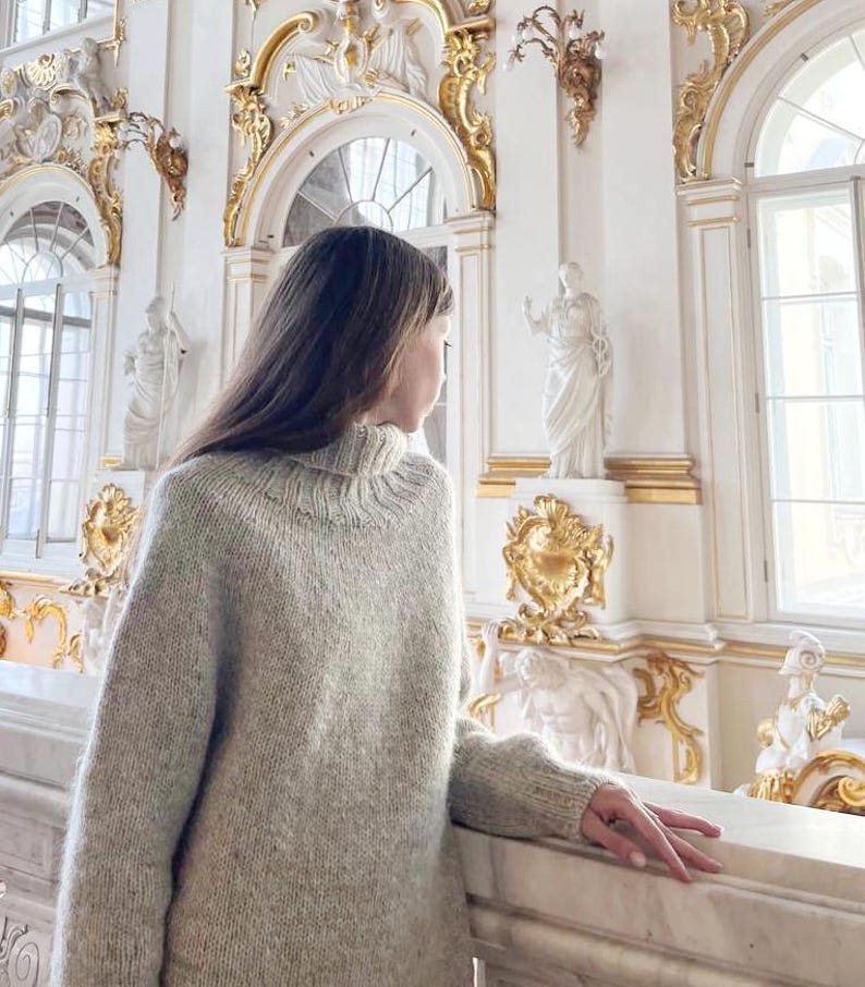 Puede incluir: Un jersey de cuello alto de color gris claro y de gran tama&ntilde;o. El jersey tiene cuello y pu&ntilde;os acanalados. La persona est&aacute; de pie en una habitaci&oacute;n con paredes blancas, detalles dorados y estatuas. La persona est&aacute; mirando por una ventana.
