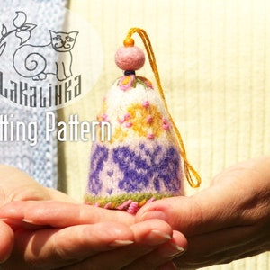 Könnte beinhalten: Ein gestrickter, rosa, weißer und violetter eiförmiger Anhänger mit einer gelben Schnur, die von oben hängt. Der Anhänger hat ein Blumenmuster und wird in den Händen einer Person gehalten. Der Text "Lakalinka Knitting Pattern" ist auf dem Bild zu sehen.