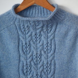 Könnte beinhalten: Hellblauer Strickpullover mit Stehkragen und Zopfmuster in der Mitte. Der Pullover hängt an einem Holzkleiderbügel. Das Strickwerk hat ein strukturiertes Aussehen.