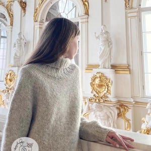 Puede incluir: Un su&eacute;ter de punto gris claro de gran tama&ntilde;o con un cuello alto acanalado. El su&eacute;ter tiene mangas largas y un corte holgado. La imagen tambi&eacute;n muestra un interior blanco y dorado ornamentado con estatuas y detalles arquitect&oacute;nicos. El su&eacute;ter es de la marca "Lakalinika".