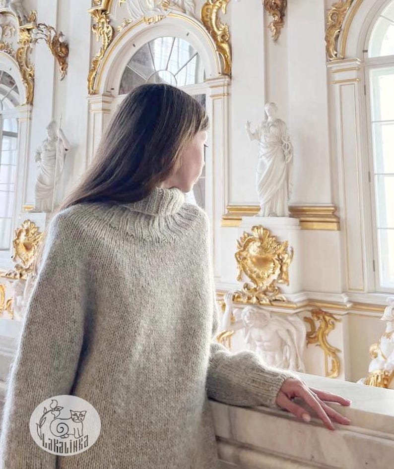 Puede incluir: Un su&eacute;ter de punto gris claro de gran tama&ntilde;o con un cuello alto acanalado. El su&eacute;ter tiene mangas largas y un corte holgado. La imagen tambi&eacute;n muestra un interior blanco y dorado ornamentado con estatuas y detalles arquitect&oacute;nicos. El su&eacute;ter es de la marca "Lakalinika".