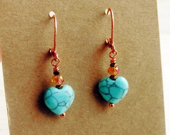 May include: A pair of turquoise heart-shaped earrings with copper-colored ear wires. Each earring features a heart-shaped bead with a black web-like pattern, accented with orange and brown beads. The earrings are displayed on a brown card.