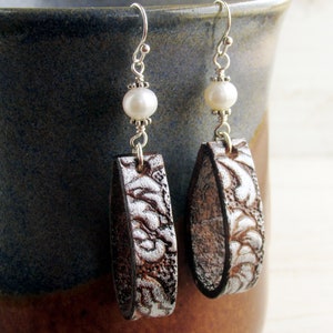 May include: A pair of handcrafted earrings featuring a silver-toned floral design on brown leather. Each earring has a white pearl bead and silver-colored ear wire. The earrings are designed to dangle.