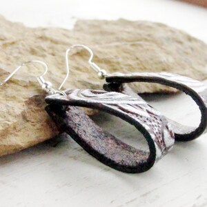 Puede incluir: Un par de pendientes ovalados hechos a mano. Los pendientes son de cuero marr&oacute;n oscuro con un dise&ntilde;o floral plateado en relieve. Los pendientes tienen ganchos para las orejas plateados.