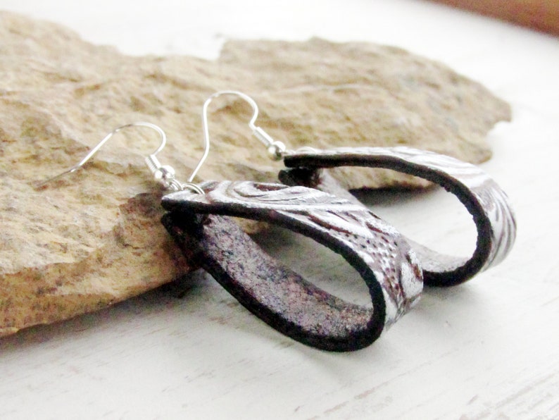 Puede incluir: Un par de pendientes ovalados hechos a mano. Los pendientes son de cuero marr&oacute;n oscuro con un dise&ntilde;o floral plateado en relieve. Los pendientes tienen ganchos para las orejas plateados.