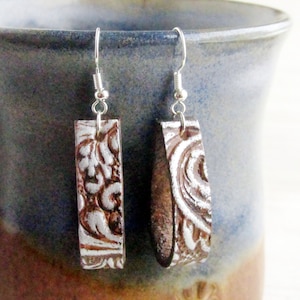Puede incluir: Un par de pendientes rectangulares con acabado plateado y dise&ntilde;o floral en relieve marr&oacute;n. Los pendientes cuelgan de ganchos plateados. Se exhiben sobre un fondo cer&aacute;mico azul y marr&oacute;n.