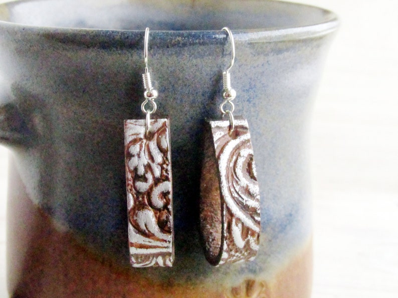 Puede incluir: Un par de pendientes rectangulares con acabado plateado y dise&ntilde;o floral en relieve marr&oacute;n. Los pendientes cuelgan de ganchos plateados. Se exhiben sobre un fondo cer&aacute;mico azul y marr&oacute;n.