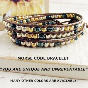 May include: Wrap bracelet with multiple strands of brown leather and beads in gold, silver, and teal. The bracelet is labeled "Morse Code Bracelet" with the text "You are unique and unrepeatable."