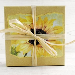 Puede incluir: Una caja de regalo cuadrada envuelta en papel marr&oacute;n claro, adornada con una imagen de girasol y atada con cinta de rafia. El girasol es amarillo con un centro oscuro, sobre un fondo azul claro y verde.