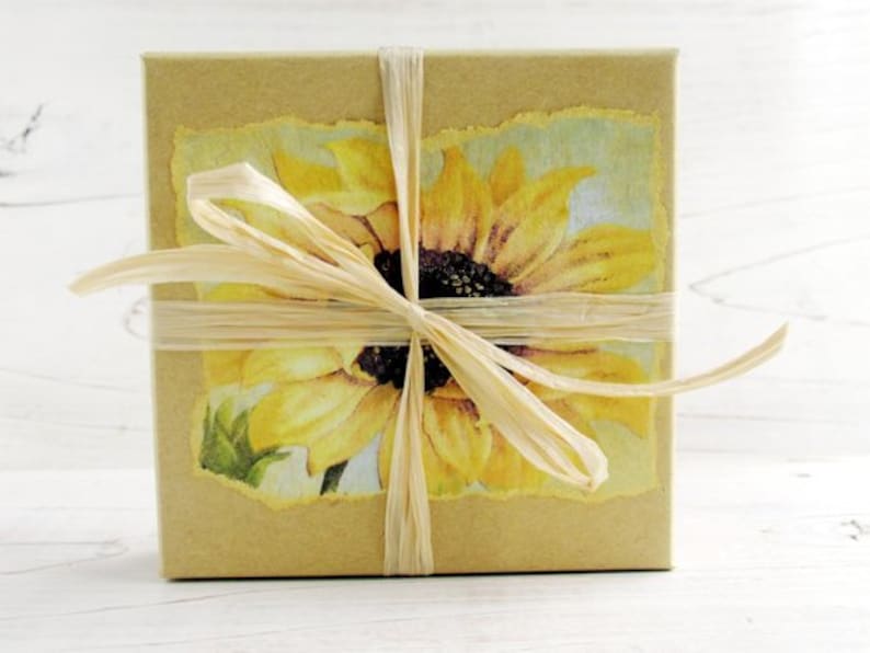 Puede incluir: Una caja de regalo cuadrada envuelta en papel marr&oacute;n claro, adornada con una imagen de girasol y atada con cinta de rafia. El girasol es amarillo con un centro oscuro, sobre un fondo azul claro y verde.