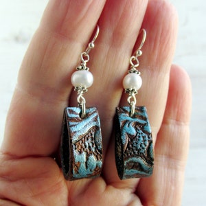 May include: Handmade earrings featuring rectangular, embossed leather pendants in brown and turquoise, accented with a white pearl and silver-tone hooks. The earrings have a rustic, bohemian style.
