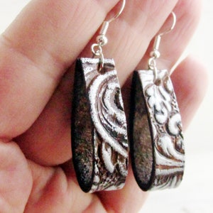 Puede incluir: Un par de pendientes rectangulares con ganchos plateados. Los pendientes tienen una base marr&oacute;n oscuro con un dise&ntilde;o plateado en relieve y ornamentado. El dise&ntilde;o parece floral o en forma de volutas. Los pendientes est&aacute;n sostenidos por una mano.