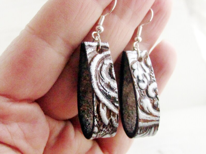 Puede incluir: Un par de pendientes rectangulares con ganchos plateados. Los pendientes tienen una base marr&oacute;n oscuro con un dise&ntilde;o plateado en relieve y ornamentado. El dise&ntilde;o parece floral o en forma de volutas. Los pendientes est&aacute;n sostenidos por una mano.
