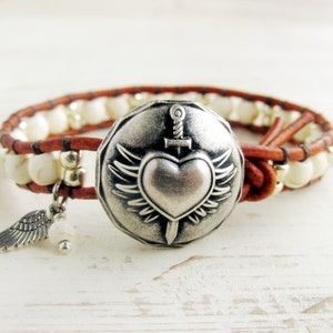 May include: Wrap bracelet featuring a silver heart and sword pendant with wings, and a small wing charm. The bracelet has a brown leather band with white beads and silver accents. A stylish accessory.