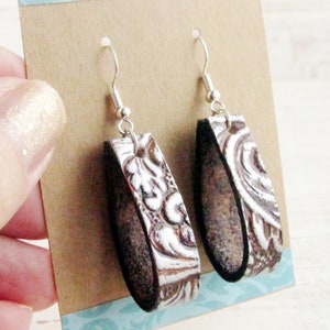 Puede incluir: Un par de pendientes colgantes con un gancho plateado. Los pendientes tienen una base de cuero marr&oacute;n con un dise&ntilde;o floral blanco en relieve. Los pendientes se muestran en una tarjeta marr&oacute;n.