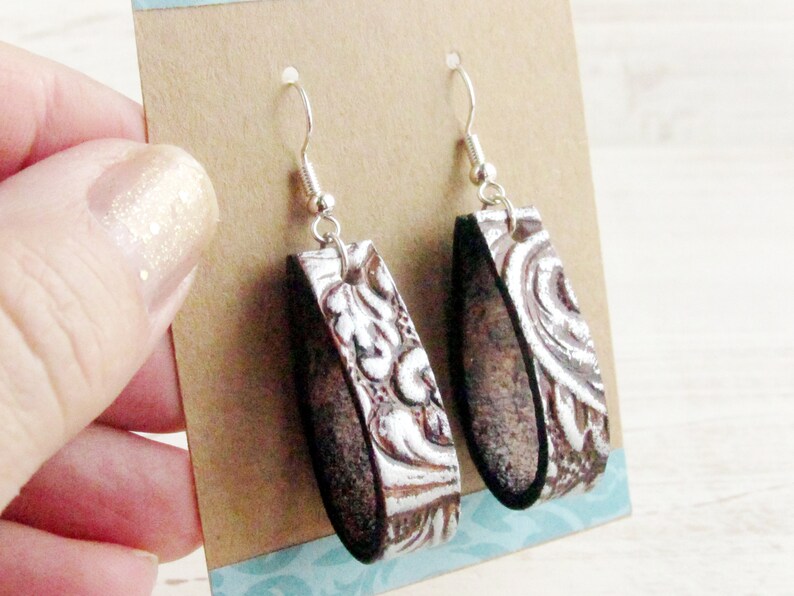 Puede incluir: Un par de pendientes colgantes con un gancho plateado. Los pendientes tienen una base de cuero marr&oacute;n con un dise&ntilde;o floral blanco en relieve. Los pendientes se muestran en una tarjeta marr&oacute;n.
