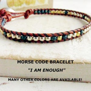 May include: A brown leather Morse code bracelet with silver and gold beads, and teal accents. The bracelet is inscribed with the phrase "I AM ENOUGH".