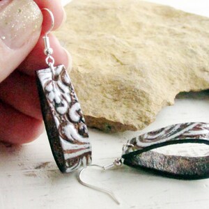 Puede incluir: Un par de pendientes rectangulares con un dise&ntilde;o en relieve marr&oacute;n y blanco. Los pendientes tienen un gancho plateado y un reverso negro. El dise&ntilde;o presenta un patr&oacute;n floral. Los pendientes se muestran sobre un fondo neutro.