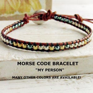 Peut inclure: Un bracelet enroulé en cuir marron orné de perles argentées, dorées et turquoise. Le bracelet est doté d'une fermeture coulissante et d'un fermoir bouton argenté. Le texte sur l'image indique "MORSE CODE BRACELET" et "MY PERSON".