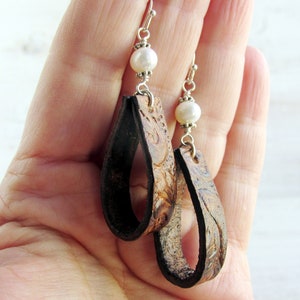 May include: A pair of handcrafted leather earrings with a teardrop design. The earrings feature a dark brown, embossed leather with a floral pattern and are accented with a white pearl bead and silver-tone metal findings.