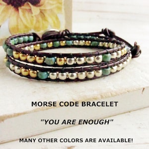 May include: A wrap bracelet with brown leather and three strands of beads. The beads are a mix of gold, silver, and turquoise. The bracelet is on a white surface. Text on the surface reads "MORSE CODE BRACELET" and "YOU ARE ENOUGH".