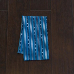 May include: A blue and white striped kitchen towel with a woven pattern. The towel is folded in half and lying flat on a dark brown wooden surface.