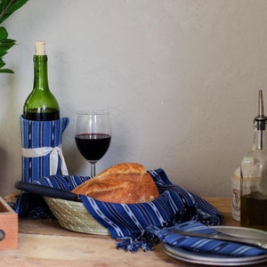 May include: A table setting with a bottle of red wine, a glass of red wine, a loaf of bread in a basket, and a blue and white striped cloth napkin. There is also a plate with a blue and white striped napkin on top of it.
