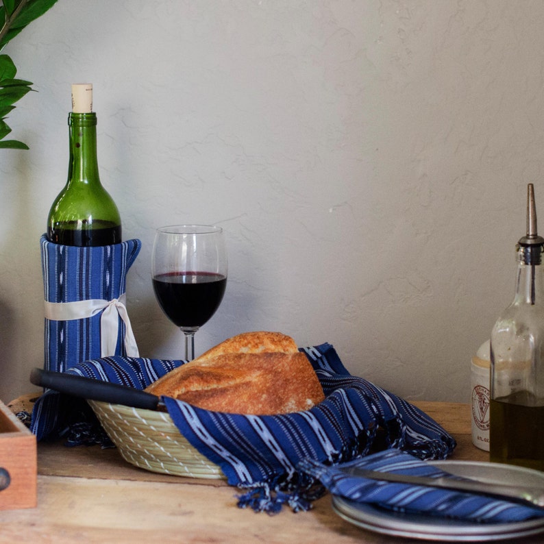 May include: A table setting with a bottle of red wine, a glass of red wine, a loaf of bread in a basket, and a blue and white striped cloth napkin. There is also a plate with a blue and white striped napkin on top of it.