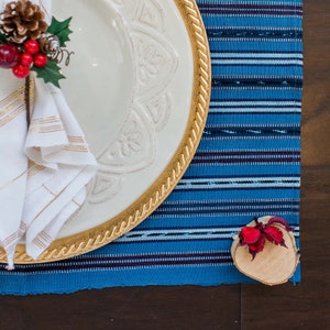 May include: A white ceramic dinner plate with a gold rim and a decorative design. The plate is set on a blue and white striped placemat with a small wooden slice with a red flower on top.