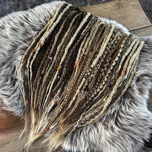 May include: A set of handmade dreadlocks in shades of brown, beige, and blonde, with decorative beads. The dreadlocks are arranged on a gray faux fur rug, showcasing various textures and styles. A warm, glowing candle is visible in the corner.