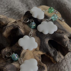 May include: White cloud-shaped charms with silver-colored findings are displayed. Each charm is connected to a small, iridescent bead. The charms are arranged on a piece of driftwood, with a soft, gray background.