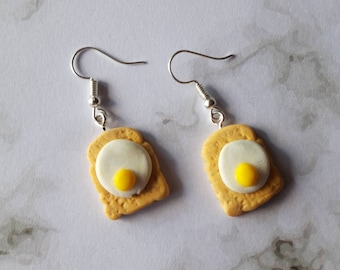 May include: A pair of earrings featuring a yellow and white fried egg on a piece of toast.