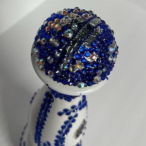 May include: Close-up view of a uniquely decorated bottle. The bottle's cap is spherical and encrusted with numerous sparkling blue and clear crystals.  A silver band around the cap is engraved with the text 