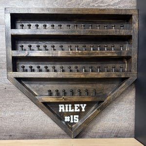 May include: A baseball-shaped wooden display case with multiple rows of pegs for holding items. The case is stained dark brown and features the name "RILEY" and the number "15" in white lettering.