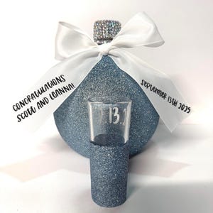 May include: A blue glitter bottle stopper with a white ribbon and a small shot glass. The ribbon reads "Congratulations Scott and Leanna!" and "September 13th 2025". The shot glass has the number "13." on it.