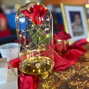 May include: A single red rose with green leaves is encased in a glass dome with a gold base. The dome is filled with white fairy lights. The rose is sitting on a red and gold sequined table runner.